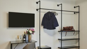 Quarto com televisão de tela plana na parede, uma escrivaninha com cadeira, uma pequena mesa com garrafa e copos, um cabideiro com jaqueta e bolsa, e uma estante.