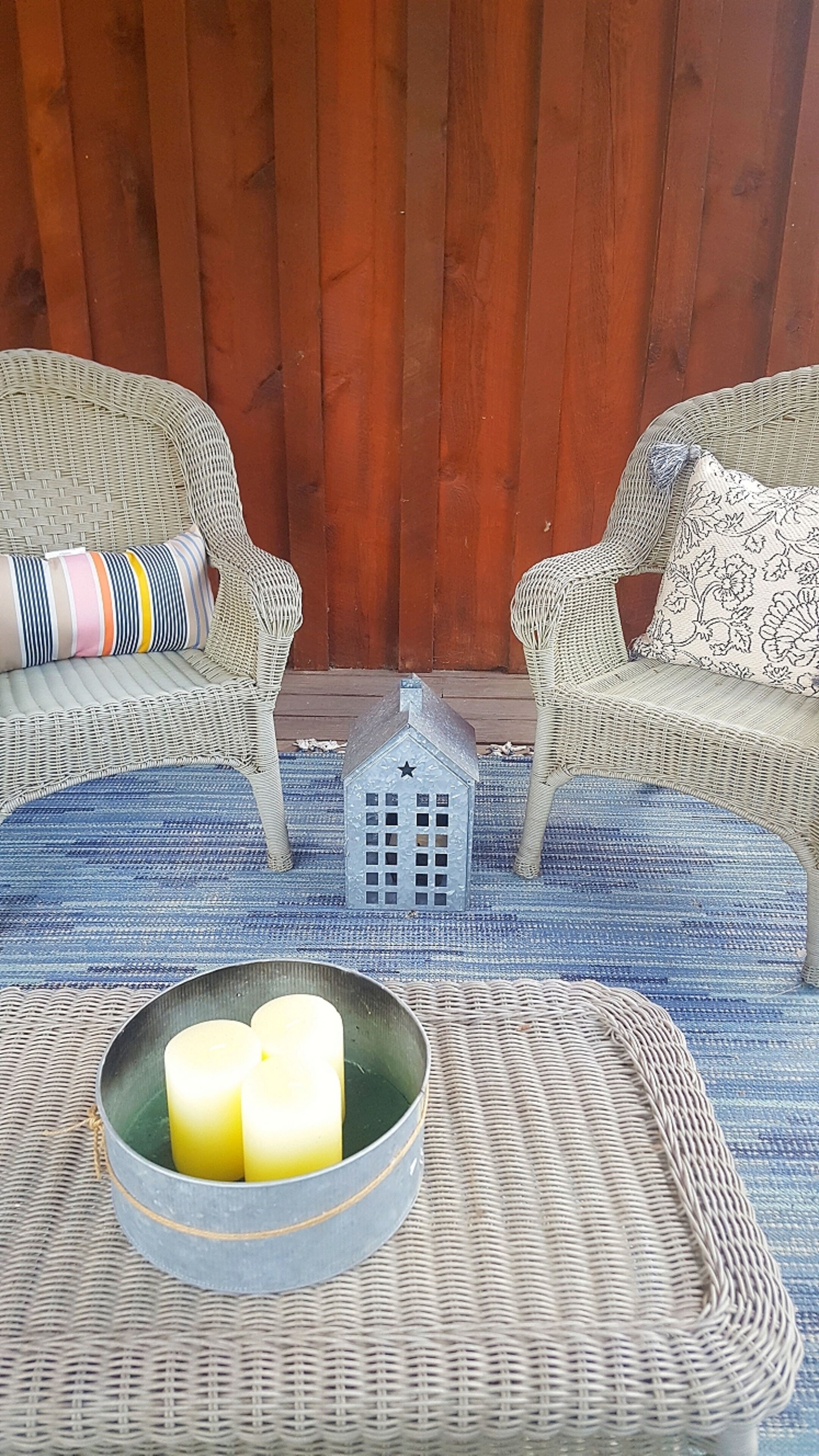 A wicker chair with a striped pillow and another with a floral pillow, a small metal house decoration, and a bowl with candles on a woven mat.