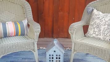 A wicker chair with a striped pillow and another with a floral pillow, a small metal house decoration, and a bowl with candles on a woven mat.