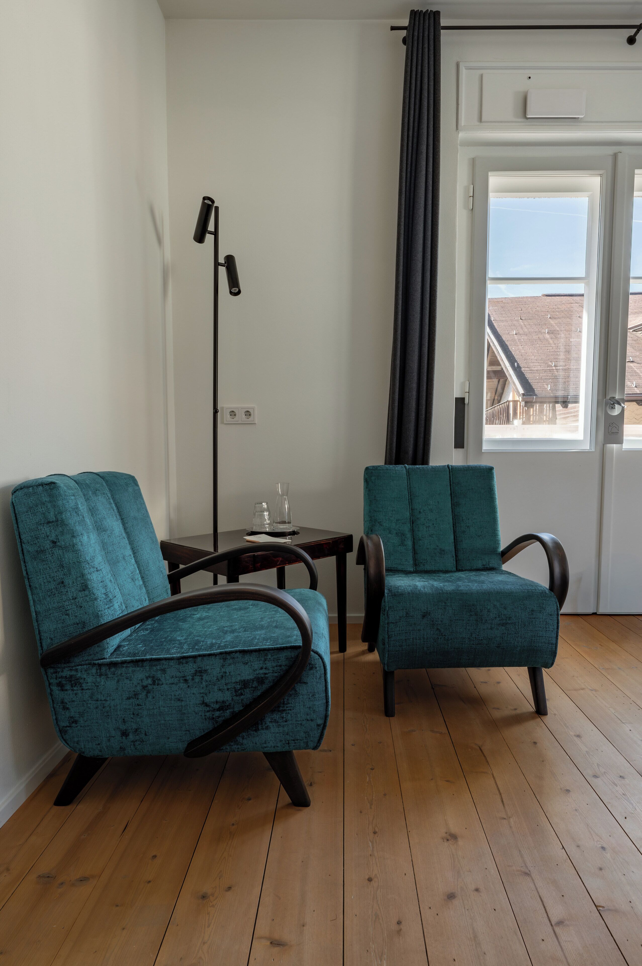 Ein Zimmer mit zwei smaragdgrünen Polstersesseln, Holzboden, einer Stehlampe, einem kleinen Tisch mit Gläsern und einem Fenster mit Blick auf ein Gebäude.