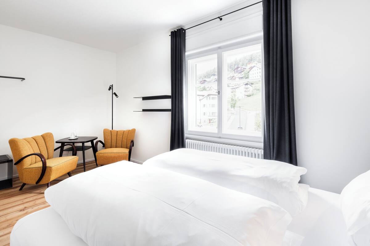Hotellihuone, jossa on suuri ikkuna, kaksi erillistä sänkyä, pieni pöytä ja kaksi keltaista nojatuolia.
