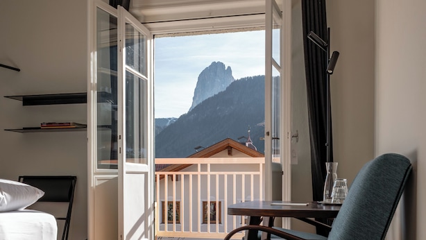 Una habitación con piso de madera, un sofá gris, una silla azul, una mesa y un balcón con vistas a la montaña.