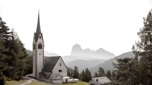 Una iglesia con una aguja alta, rodeada de árboles y montañas al fondo.