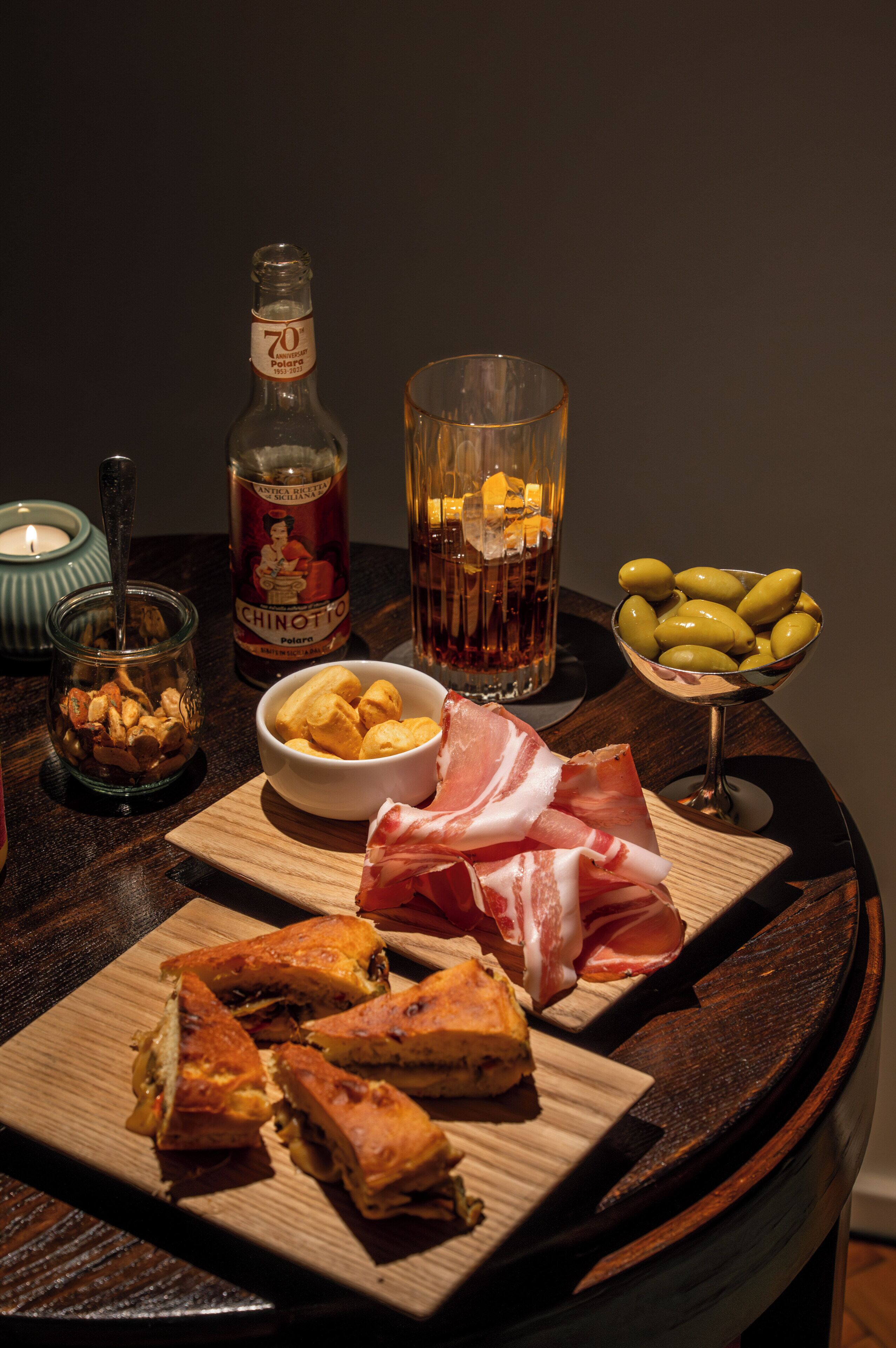 A wooden table with a bottle of Chinotto, a glass of whiskey, olives, a bowl of nuts, and a plate of sandwiches.