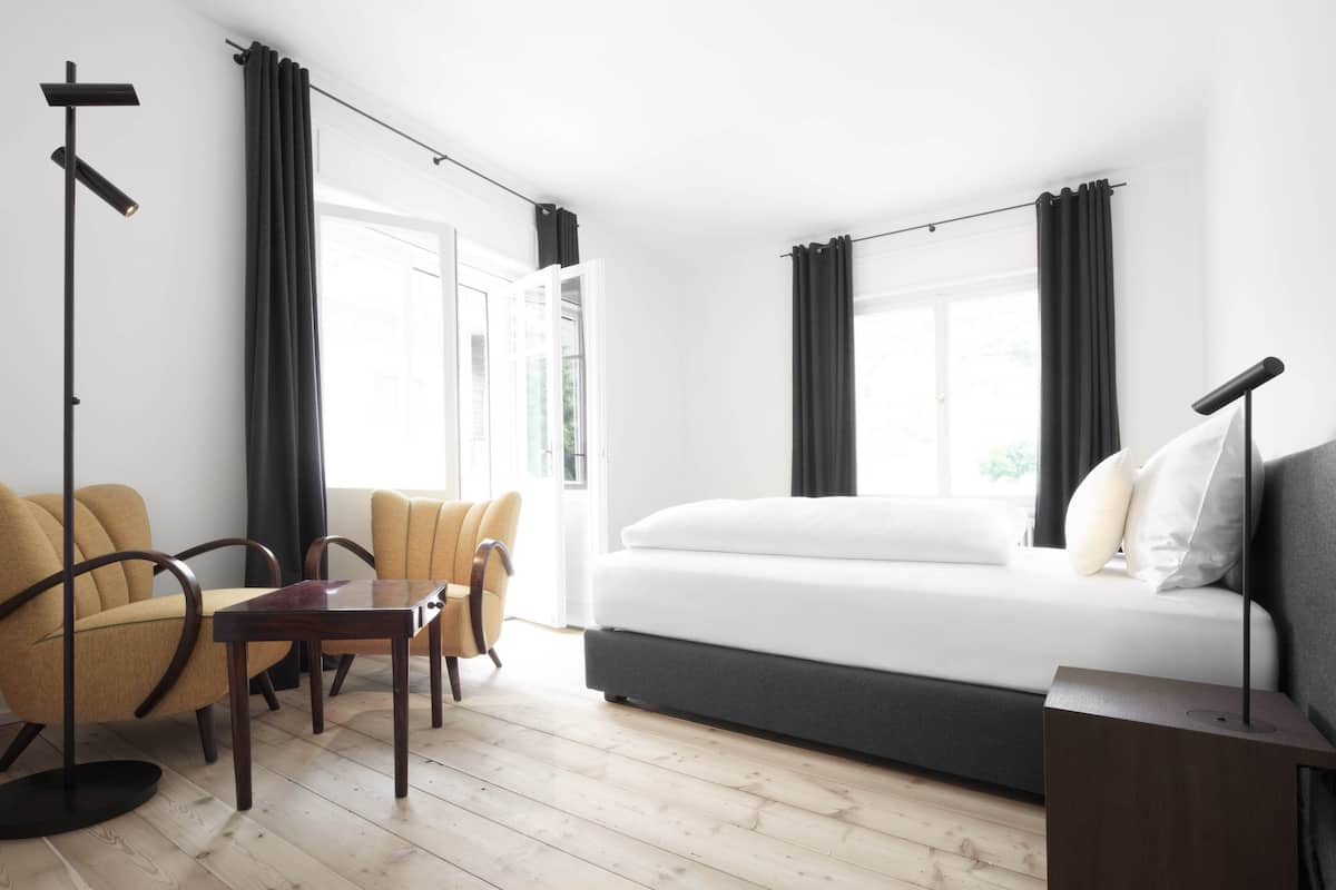 Moderni hotellihuone, jossa on sänky, työpöytä, kaksi tuolia ja lattiavalaisin.