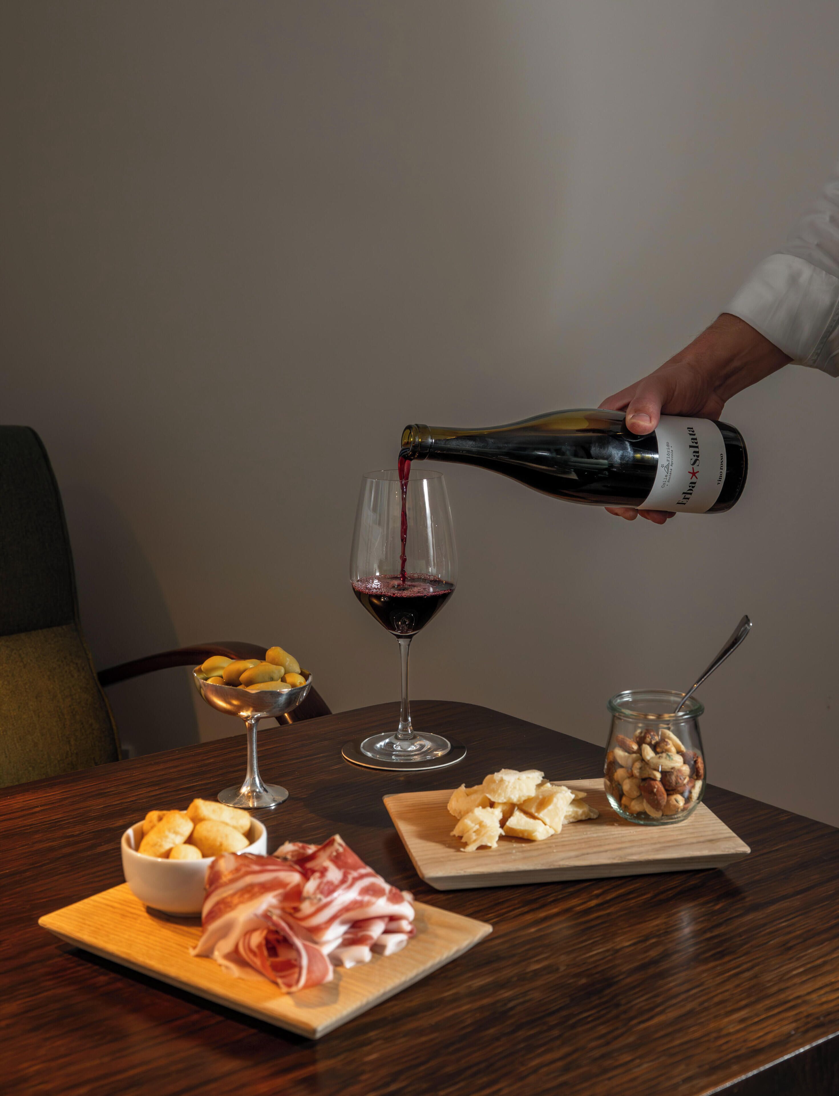Uma mesa com uma garrafa de vinho sendo servida em um copo, uma cesta de pão, um pote de nozes e um prato de carne curada.
