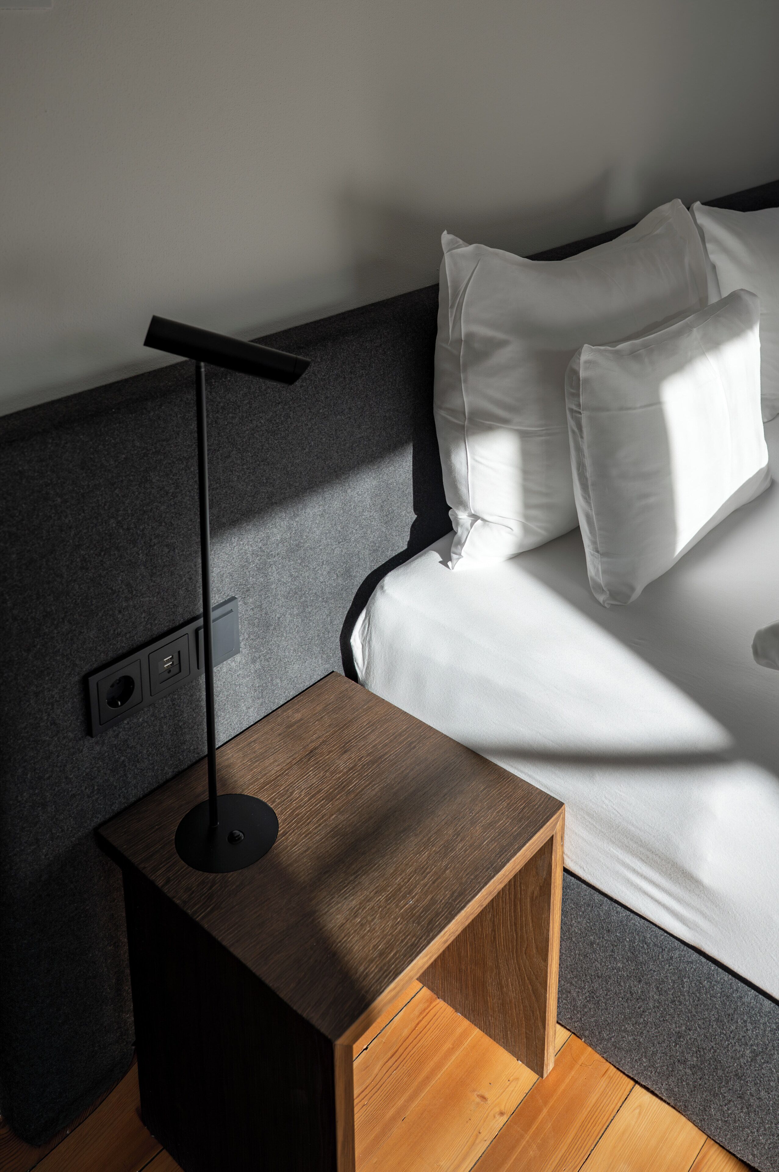 A modern bedroom with a wooden nightstand, a black floor lamp, a bed with white bedding, and a grey headboard.