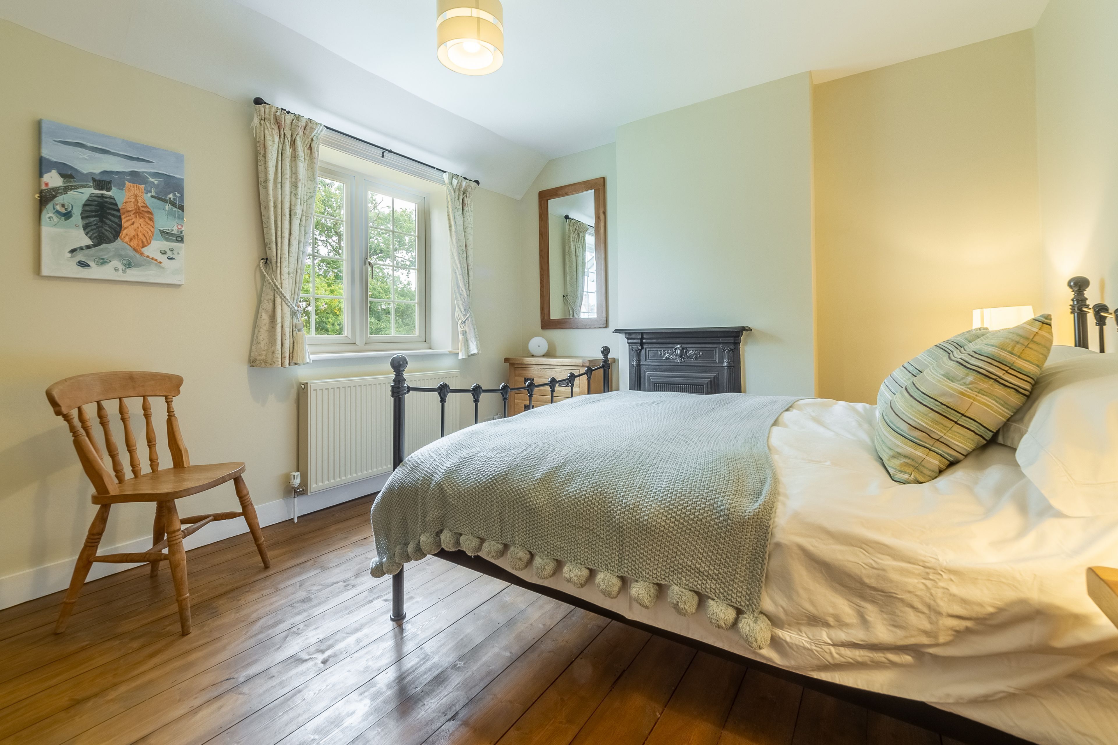 A bedroom with a wooden floor, a bed with a white cover, a chair, a window with curtains, a radiator, a mirror, and a painting on the wall.