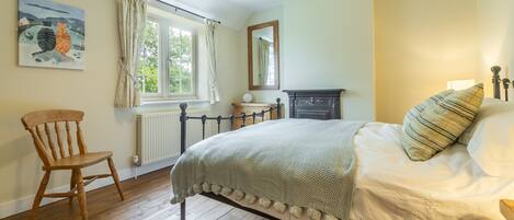 A bedroom with a wooden floor, a bed with a white cover, a chair, a window with curtains, a radiator, a mirror, and a painting on the wall.