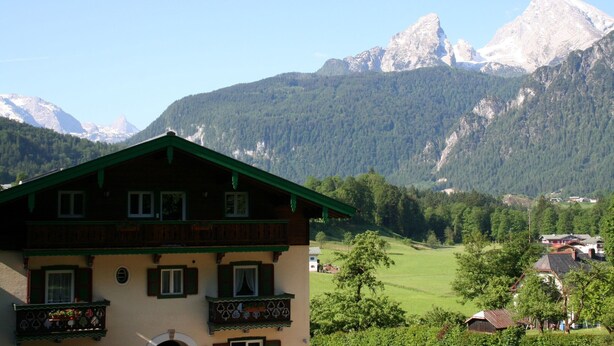Ein traditionelles Chalet mit einem grünen Dach und einem Balkon, vor einer Berglandschaft und blauem Himmel.