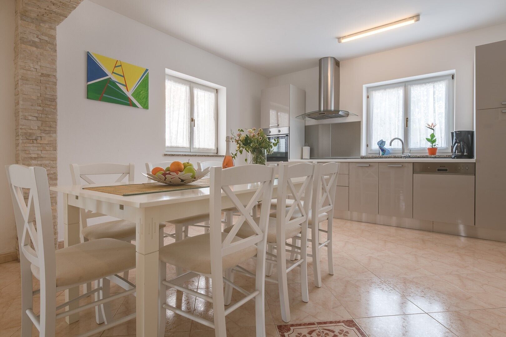 A modern kitchen with white cabinetry, a stainless steel range hood, and a dining area with white chairs and a table set with fruit.