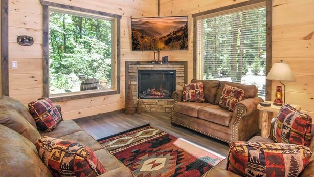 Interior acogedor de una cabaña con chimenea, paredes de madera, un conjunto de sofás y ventanas amplias.