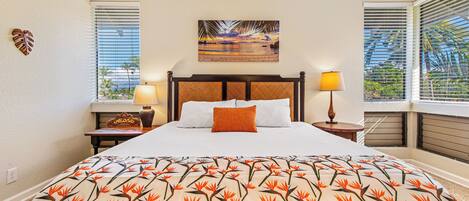 Une chambre d’hôtel avec un lit, une table de chevet, une fenêtre avec des stores, un tableau au mur et une couvre-lit aux motifs tropicaux.