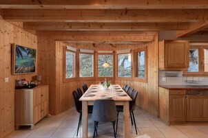 A wooden-paneled dining room with a large table set for eight, flanked by chairs, and a kitchen area with wooden cabinets and a window.