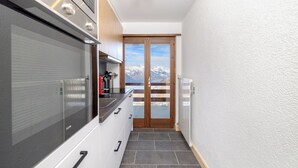 A narrow kitchen with a large oven, a window with a mountain view, and a door leading to a balcony. - App Les Rocailles 9 Family Retreat With Sauna (Riddes)