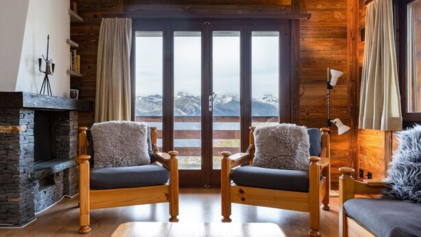 A cozy living room with a stone fireplace, two wooden chairs with fluffy cushions, and a view of snow-capped mountains through large glass doors. - App Les Rocailles 9 Family Retreat With Sauna (Riddes)