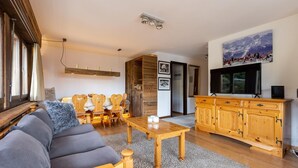 A cozy living room with a sofa, wooden dining table, and a TV. - App Les Rocailles 9 Family Retreat With Sauna (Riddes)