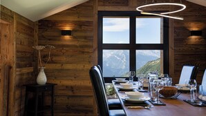 A dining area with a wooden interior, a large window offering a mountain view, and a modern pendant light. - Chalet Lupin (Riddes)