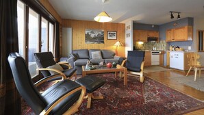 Una sala de estar acogedora con un sofá, sillas, una mesa de centro y una alfombra. Cuenta con una zona de cocina con armarios de madera y un refrigerador.