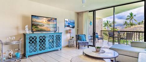 A spacious living area with a large TV, a blue cabinet, a round table, and a balcony with a view of palm trees.