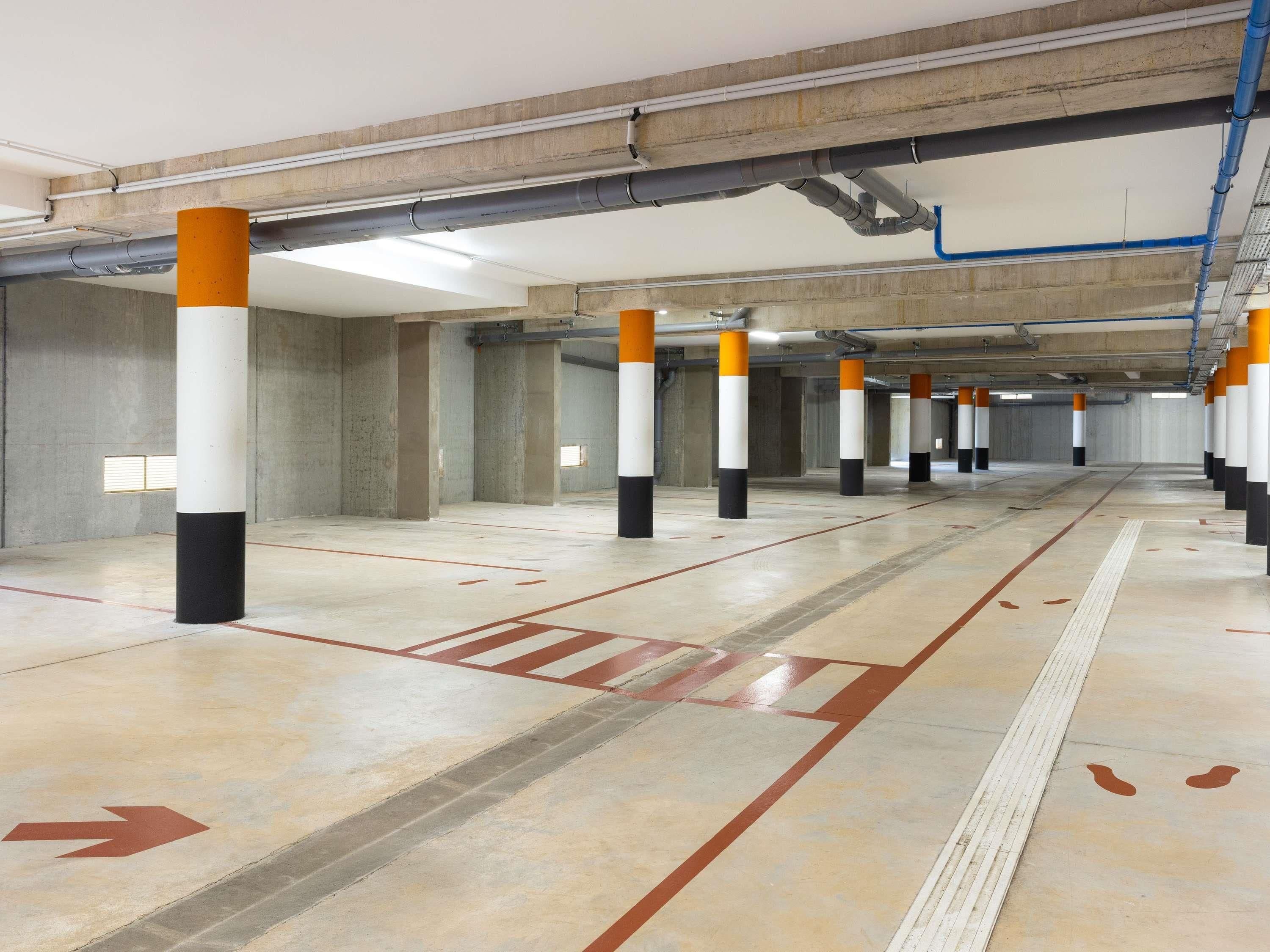 A parking garage with concrete pillars, directional arrows on the floor, and overhead pipes.