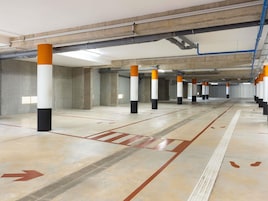 Un garaje de estacionamiento con pilares de concreto, flechas direccionales en el suelo y tuberías elevadas.