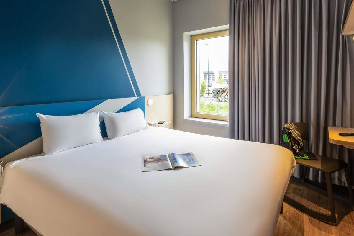 Ett hotellrum med en stor säng, ett fönster med gardiner, ett litet bord och en stol.