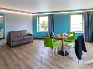 A modern room with a sofa, a dining table with chairs, and large windows. - ibis budget Épinal (Epinal)