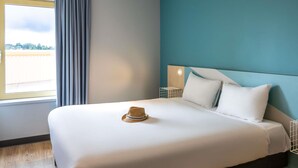 Uma cama bem arrumada com um chapéu, uma mesa de cabeceira com abajur e vista para o exterior pela janela.