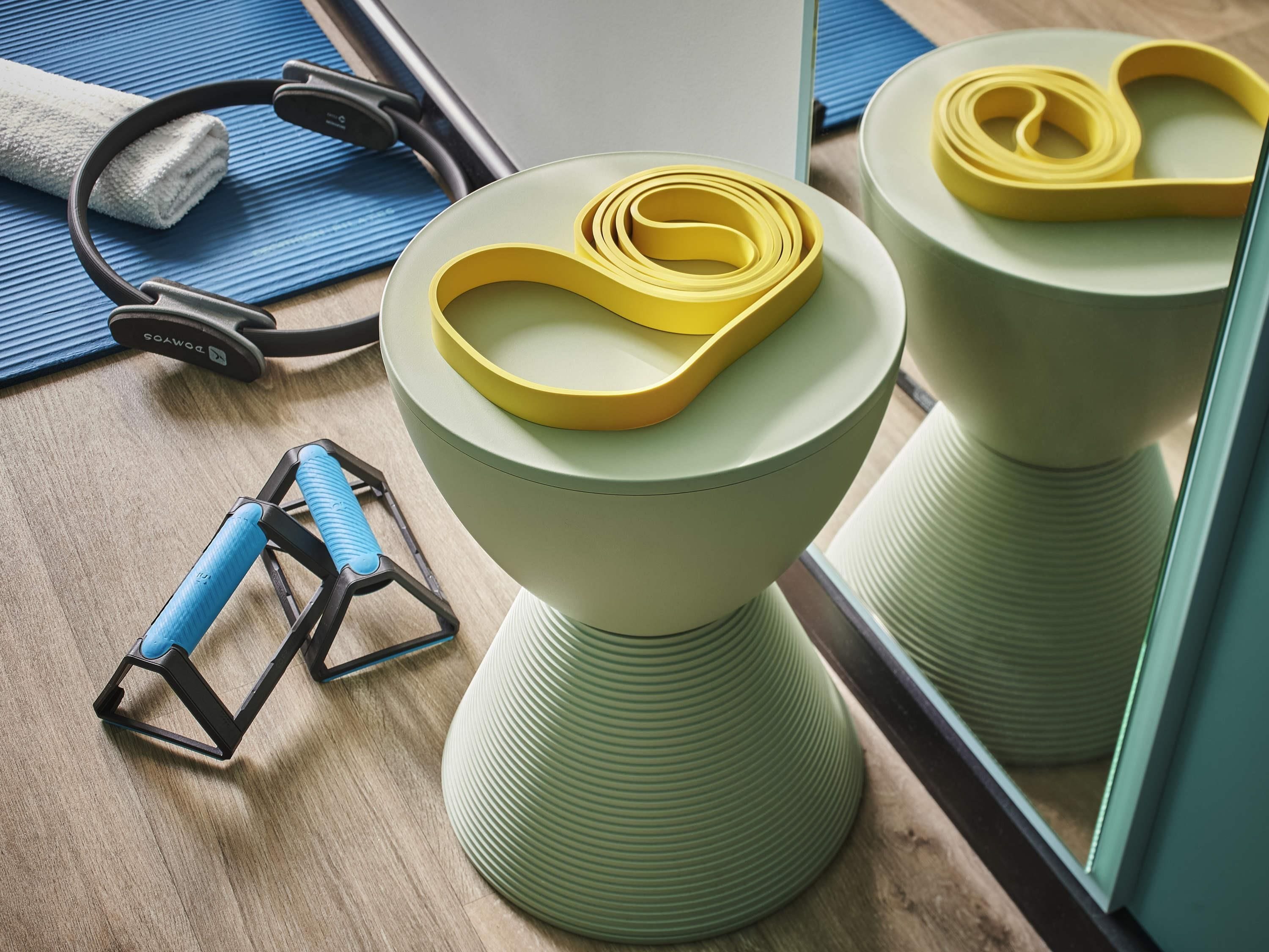 A room with a wooden floor, a blue exercise mat, a pair of headphones, a yellow resistance band, and a mirror reflecting the room.