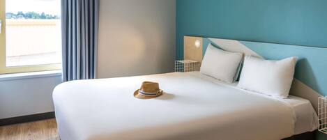 Un lit bien fait, sur lequel repose un chapeau, une table de chevet avec une lampe, et une vue sur l’extérieur par une fenêtre.