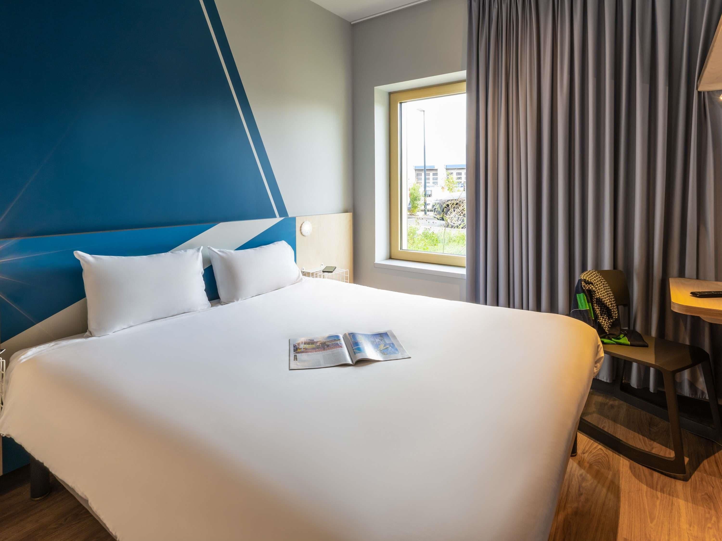 A hotel room with a large bed, a window with curtains, a small table, and a chair.