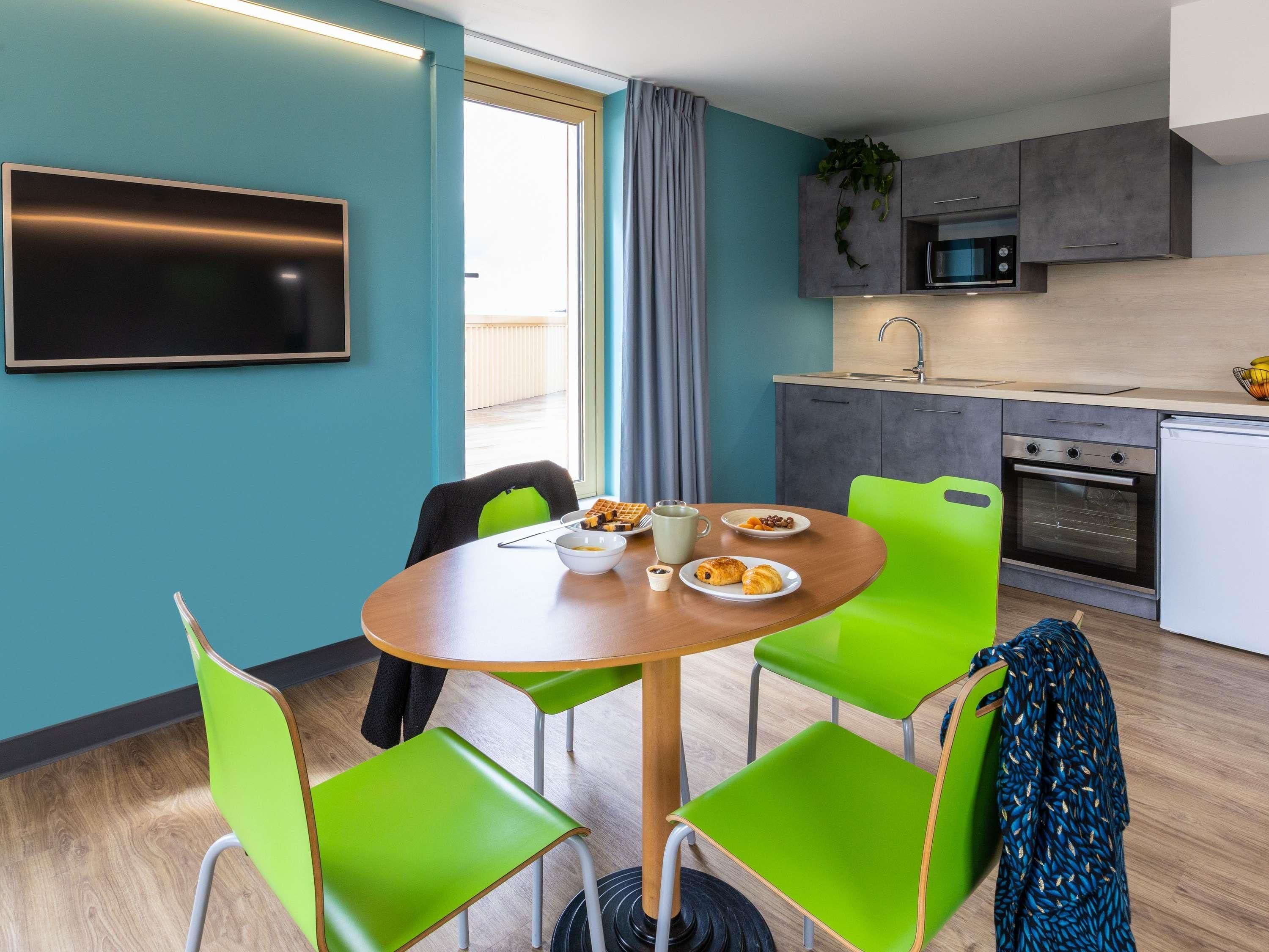 A modern kitchen with a microwave, oven, and sink. There is a dining table with chairs and a TV mounted on the wall.