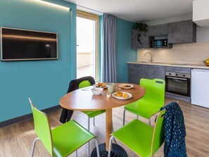 A modern kitchen with a microwave, oven, and sink. There is a dining table with chairs and a TV mounted on the wall. - ibis budget Épinal (Epinal)