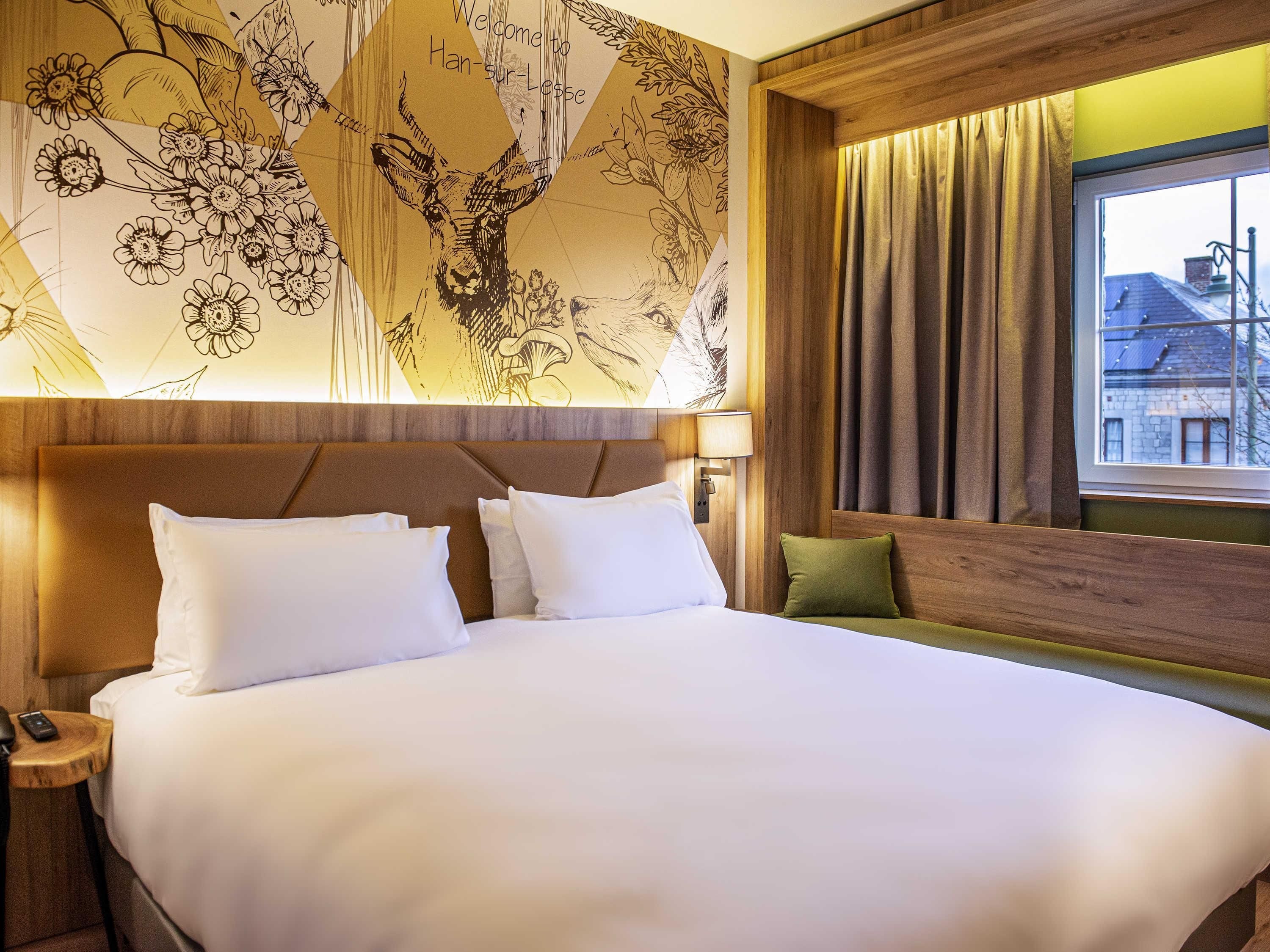 A hotel room with a large bed, a nightstand, a lamp, a window with curtains, and a wall with a floral pattern.
