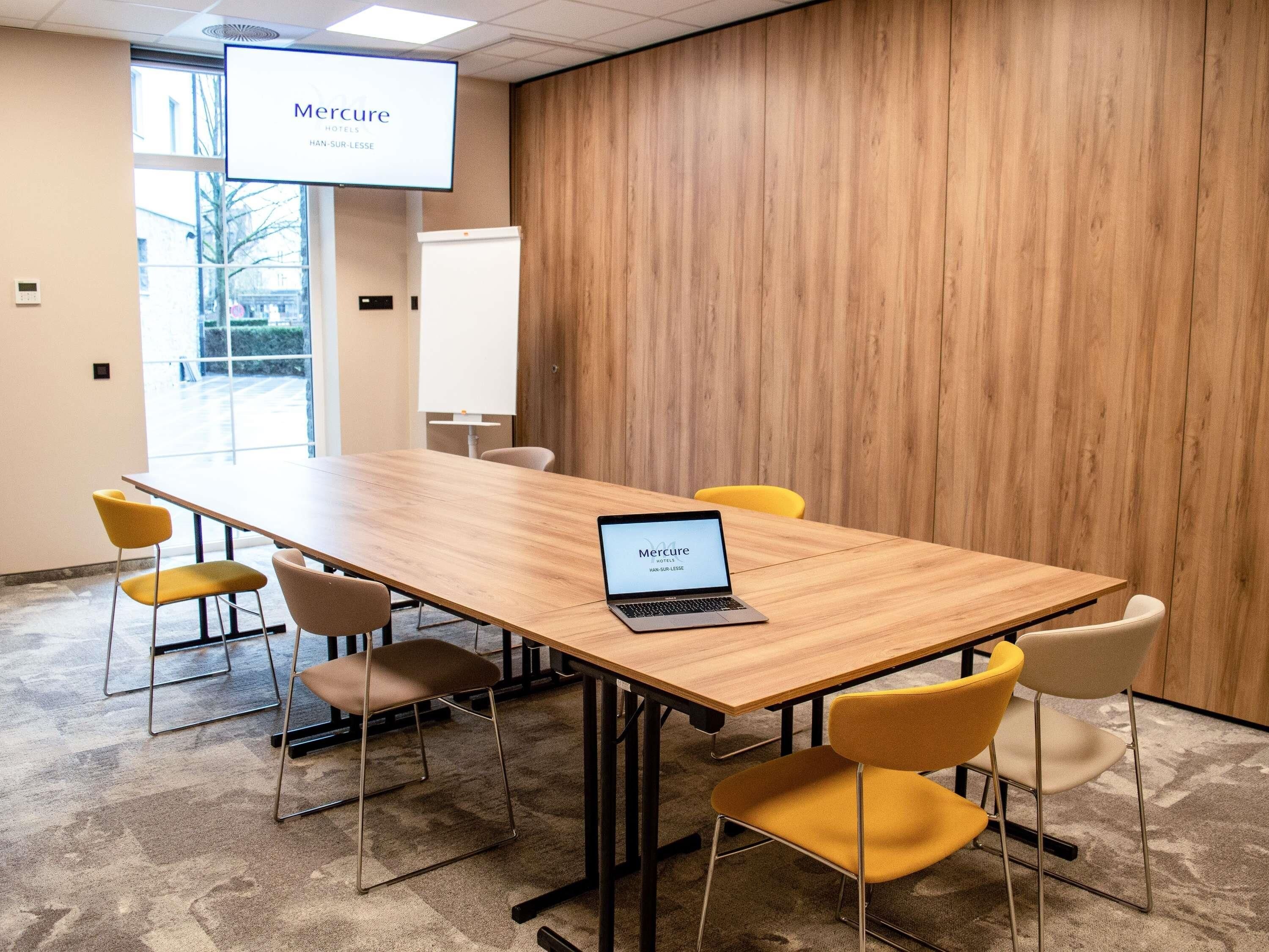 Een vergaderruimte met een houten tafel, gele en witte stoelen, een laptop en een scherm waarop 'Mercure Hotels' staat.
