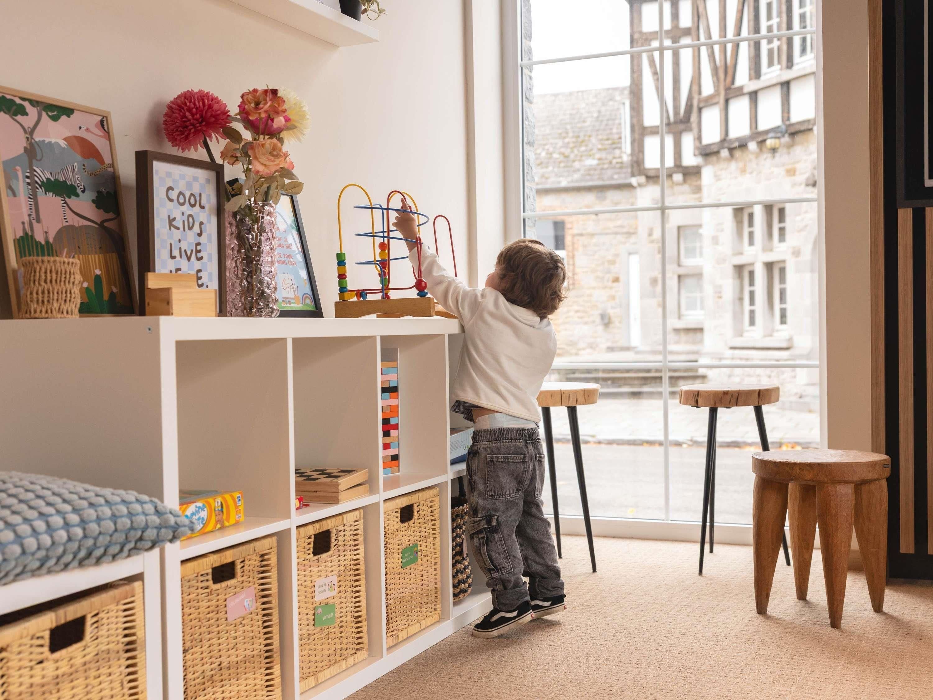 A child playing with a toy at a table in a room with large windows, wicker baskets, and a shelf with framed pictures.