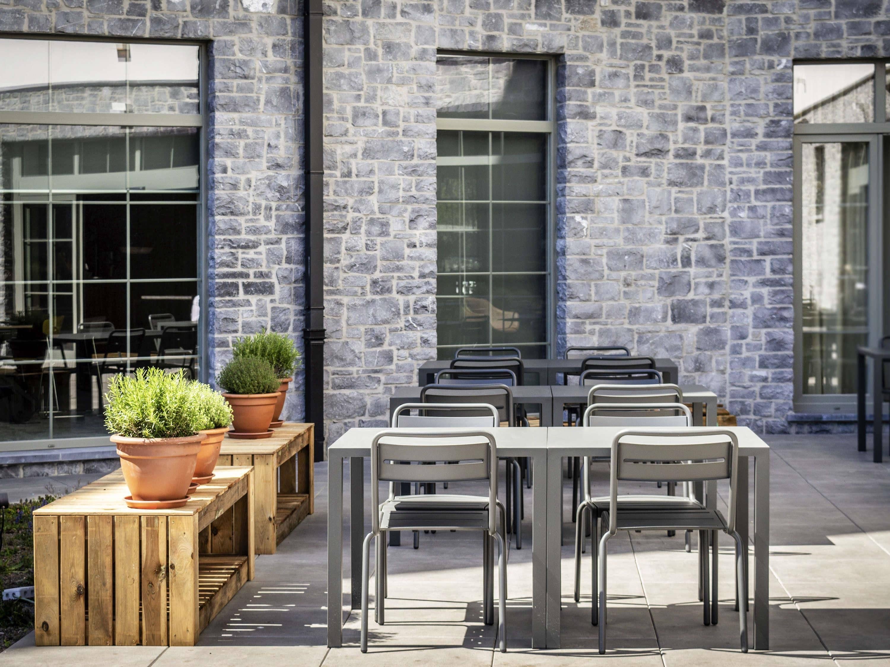 An outdoor seating area with a stone wall, wooden tables, metal chairs, and potted plants.