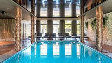 Une piscine intérieure avec de grandes fenêtres offrant une vue dégagée sur le ciel, entourée de chaises longues et de piliers décoratifs.