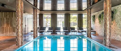 An indoor swimming pool with a clear view of the sky through large windows, surrounded by lounge chairs and decorative pillars.