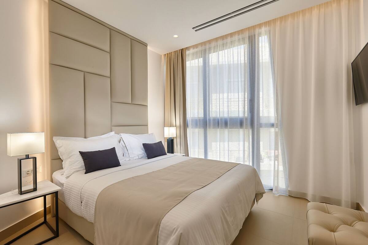A modern hotel room with a large bed, bedside lamps, a flat-screen TV, and a view of the city through sheer curtains.