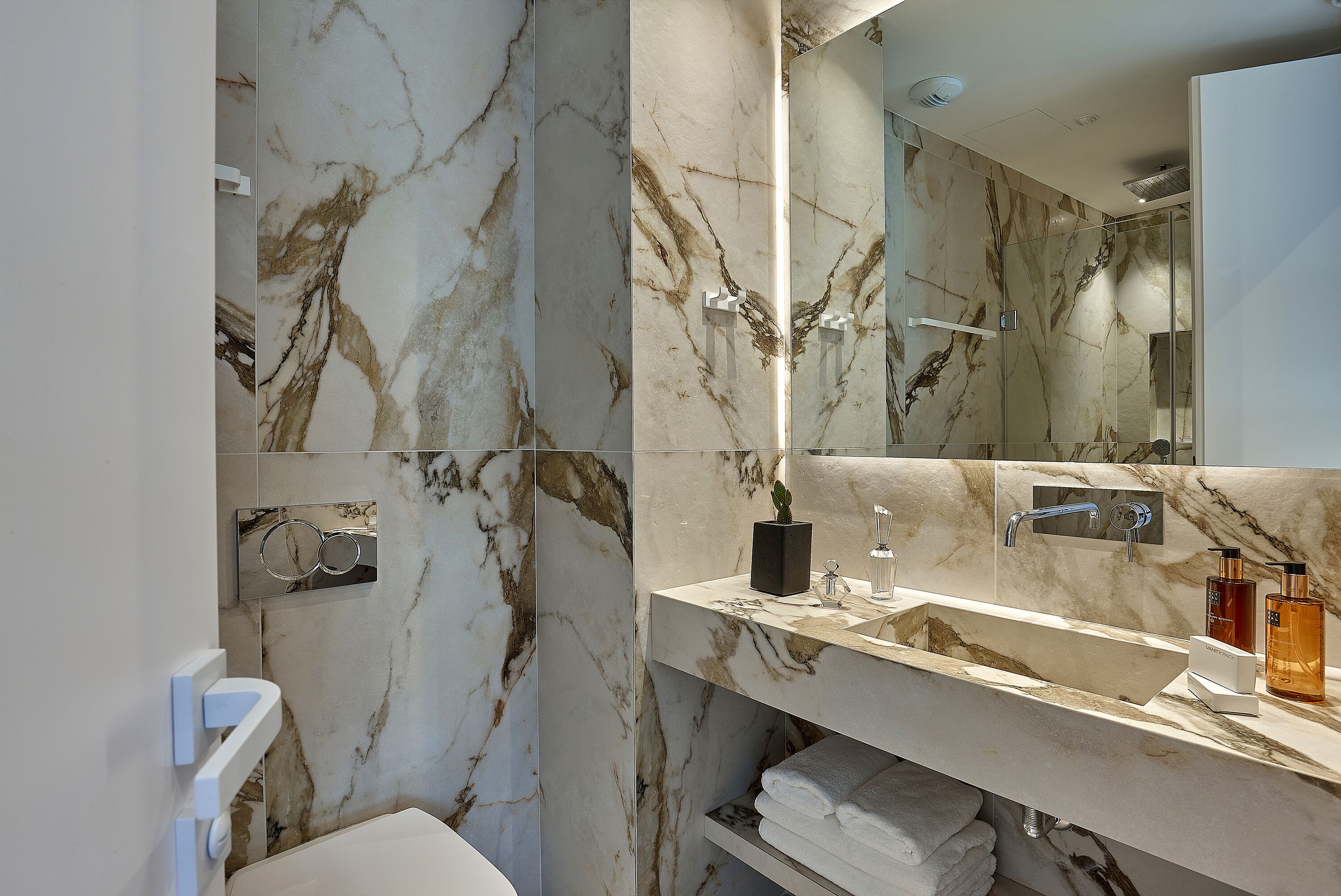 Un baño moderno con paredes de mármol, un espejo grande y un mueble de tocador con artículos de aseo.