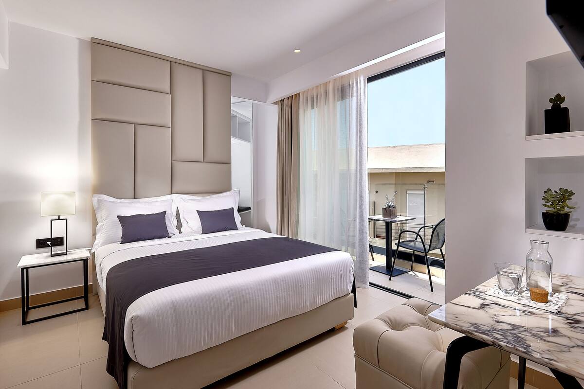 A modern hotel room with a large bed, a small table with a vase, a chair, and a balcony with a table and chairs.