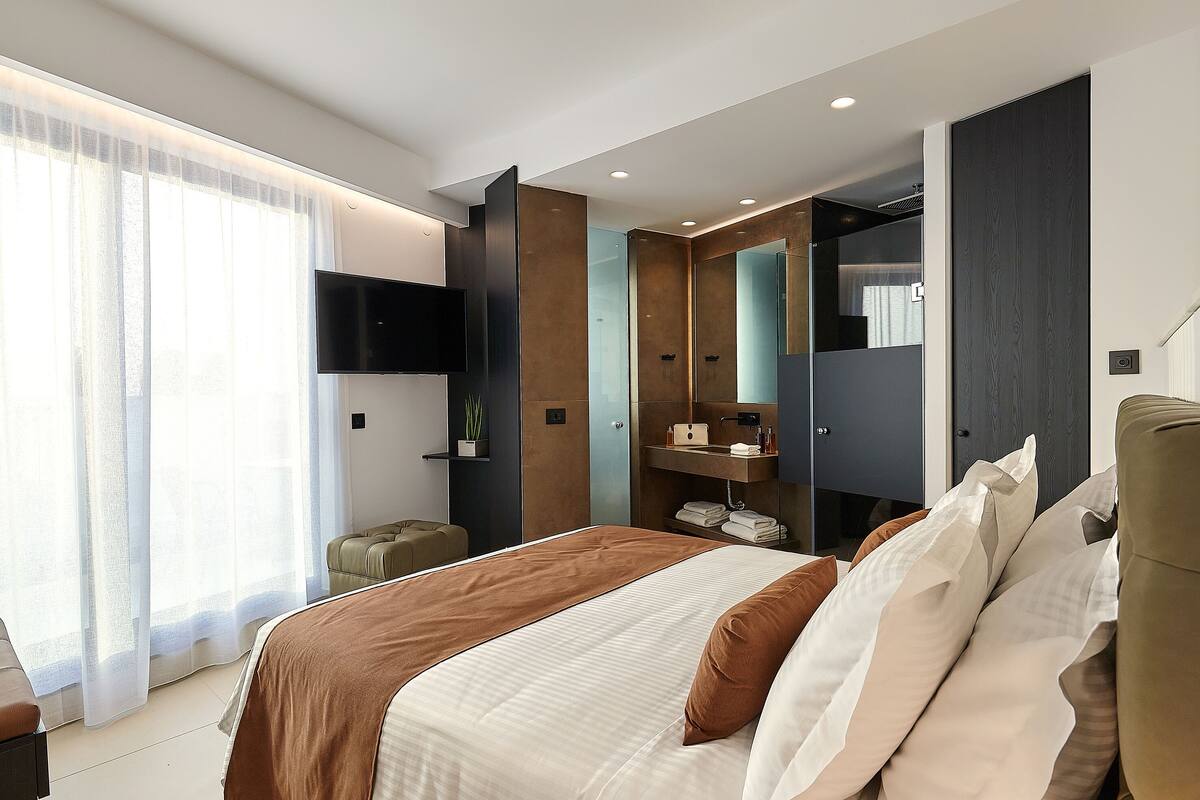 A modern hotel room with a large bed, a TV mounted on the wall, a wardrobe, and a bathroom visible through a glass door.