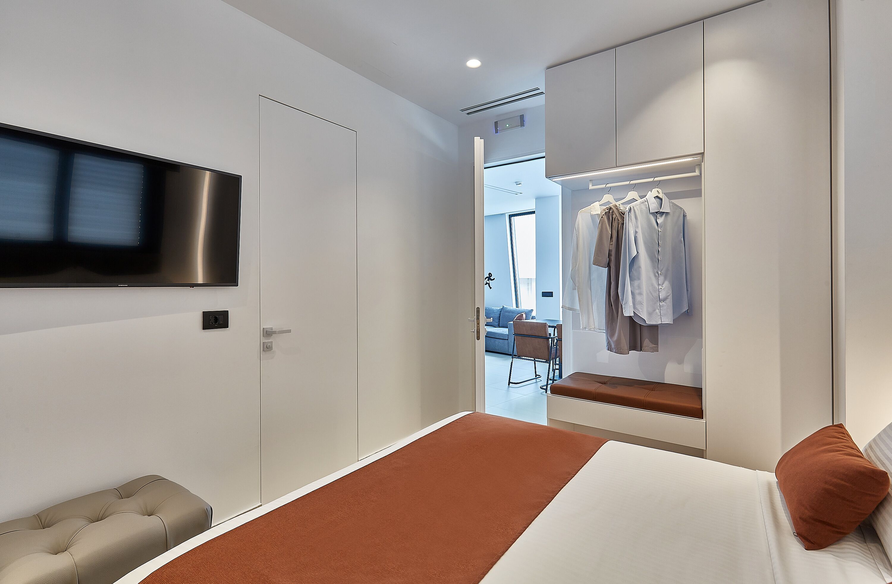 A modern bedroom with a bed, a TV mounted on the wall, a wardrobe, and a seating area with a bench.