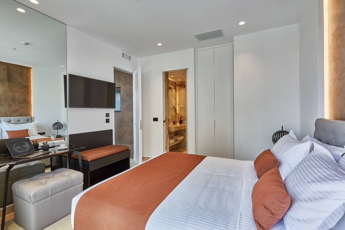 A modern hotel room with a large bed, a desk with a laptop, a seating area, and a walk-in closet.