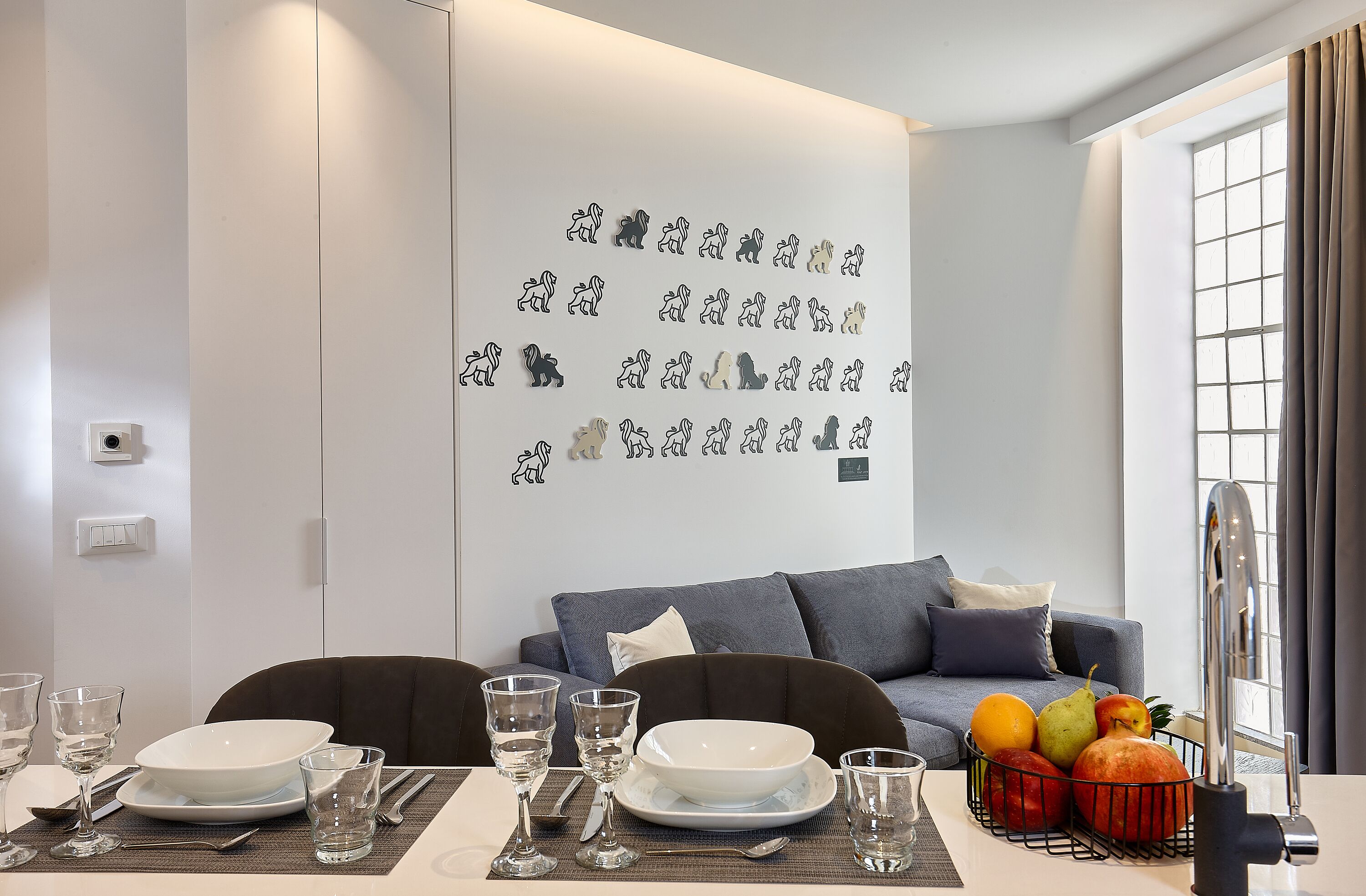 A modern dining area with a white table set for two, a blue sofa, and a fruit basket on the table.