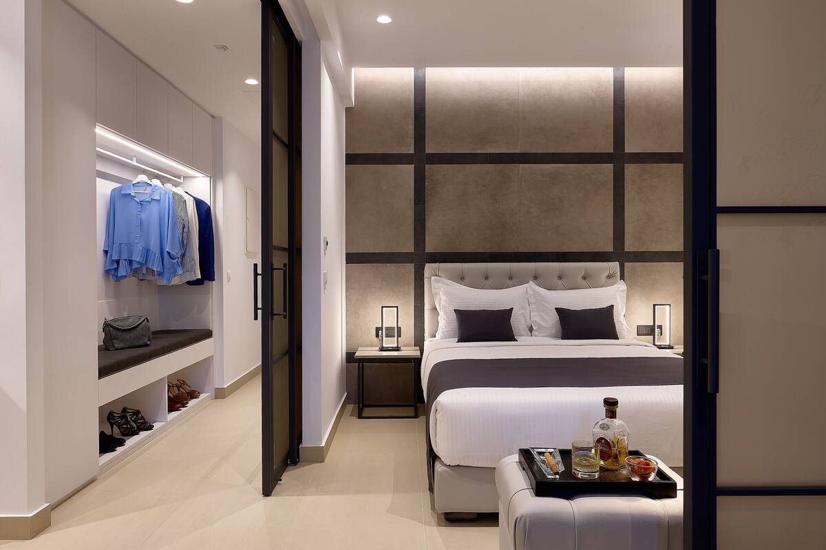 A modern hotel room with a large bed, bedside tables, a wardrobe, and a seating area with a tray of drinks.