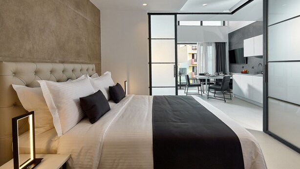 Um quarto moderno com uma cama grande, uma mesa de cabeceira com abajur, e vista para a área da cozinha adjacente.