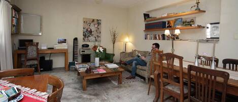 Sala de estar com sofá, mesa de centro e cadeiras, uma escrivaninha com computador e uma estante com livros e objetos decorativos.